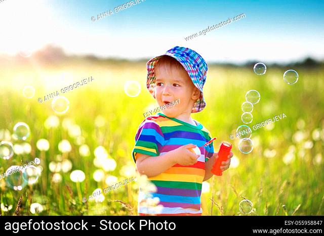 Happy baby boy standing in grass on the field with dandelions at sunny summer evening. child outdoors in nature at sunset blowing bubbles