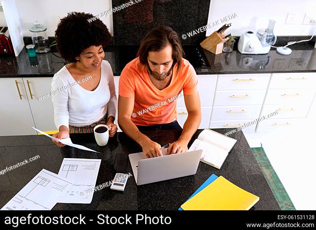 Diverse couple sitting in kitchen using laptop and paying bills