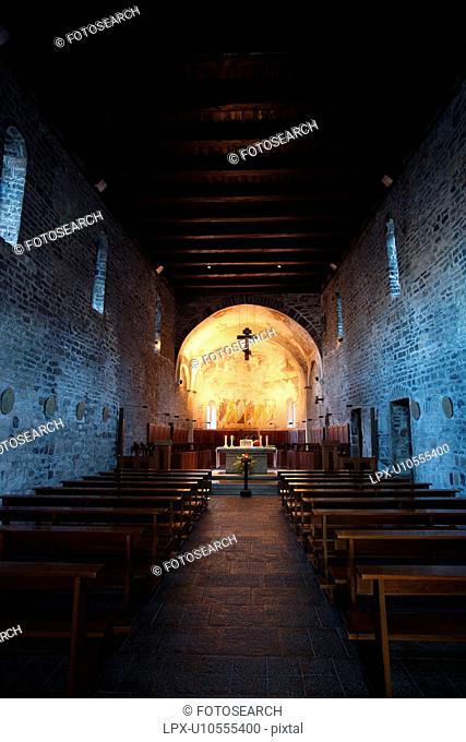 Interior view of nave of Abbey, Piona, Lake Como, Northern Italy, showing stone walls and flat wooden beam ceiling, with altar illuminated, and frescoed apse