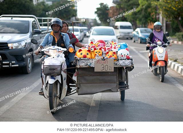 19 October 2019, Thailand, Chiang Mai: A street hawker sits on a motorcycle next to his goods and waits at a traffic light in traffic