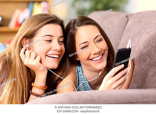 Front view portrait of two joyful friends listening music on line from a smart phone lying on a couch in the living room at home