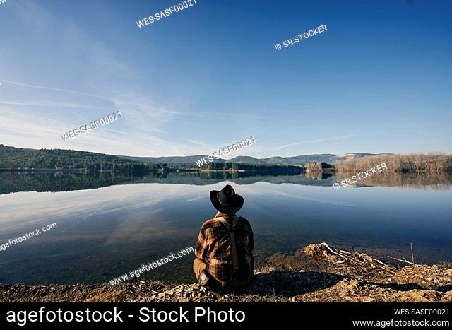 Bushcrafter looking at lake while sitting under blue sky