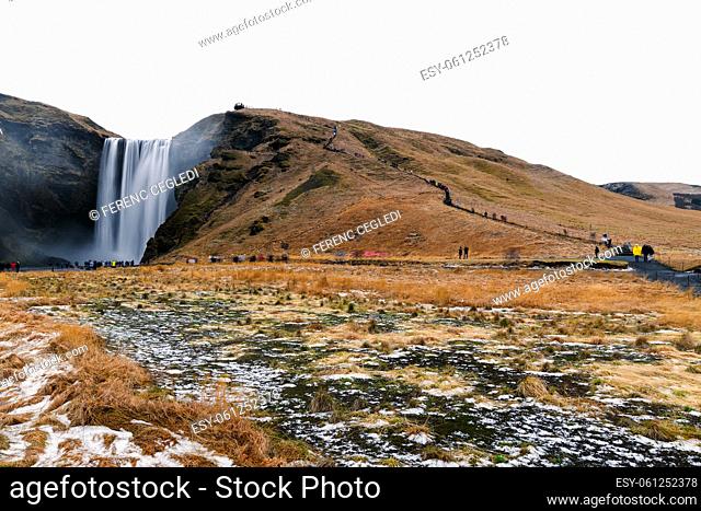 The Iconic Skogafoss Waterfall In Iceland. There are unrecognizable tourist people with motion blur in the foreground