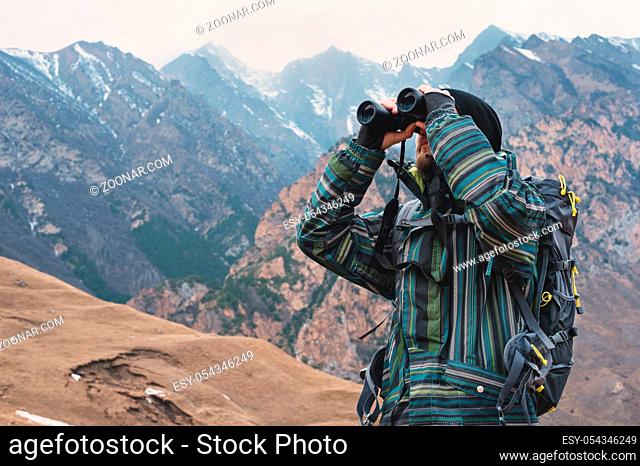 A hipster man with a beard in a hat, a jacket, and a backpack in the caucasian mountains holds binoculars, adventure, tourism, tracking