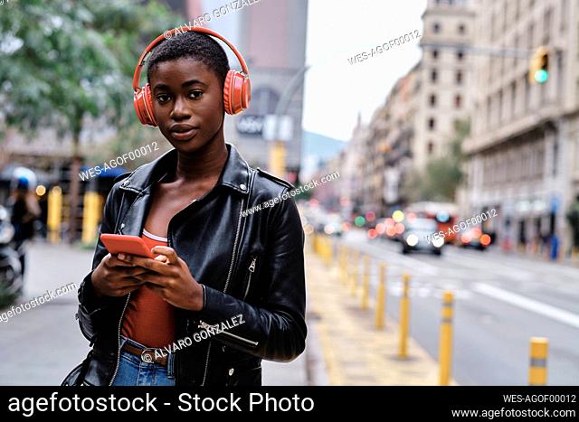 Woman wearing headphones and jacket using smart phone while standing in city