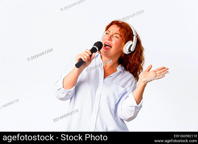 Emotions, lifestyle and leisure concept. Carefree pretty middle-aged redhead woman in headphones, holding microphone and singing, playing karaoke game