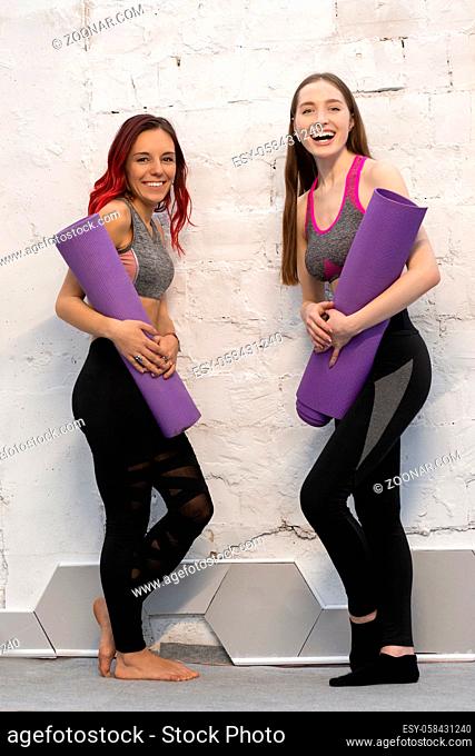 Two pretty fitness girls in sports out fits holding a yoga mat standing next to the white wall, smiling on camera isolated on wall background