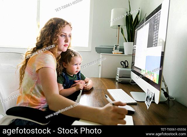 Young mother with her daughter on lap working from home at desk