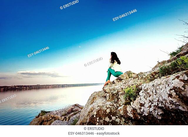 Young woman sits on the rock and enjoys the view of the river at sunset
