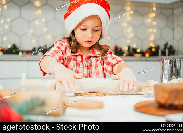 Little dark-haired girl 3 years old in red Christmas cap and checkered shirt roll out dough for gingerbread cookies in white Christmas decorated kitchen with...
