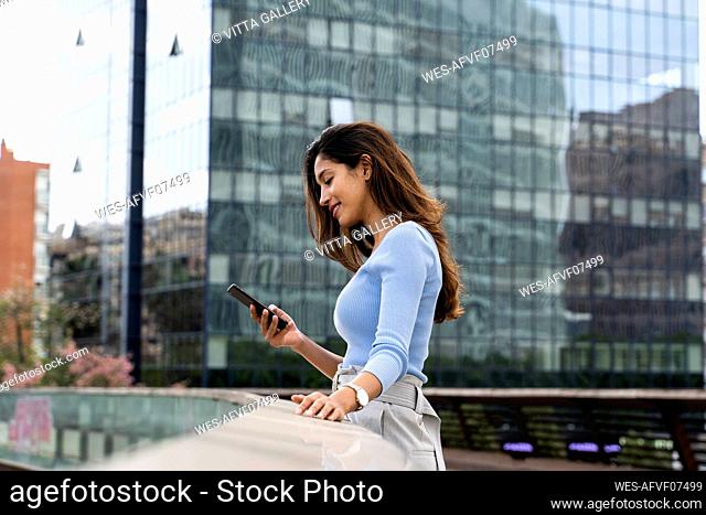 Smiling young businesswoman using smart phone while standing on footbridge