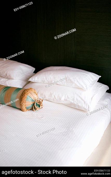 A bed with a white blanket and pillows of different sizes and an elongated cushion in a room with dark walls. High quality photo