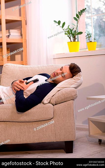 Lazy man being asleep at his home on sofa. Young male holding TV remote while sleeping in living room