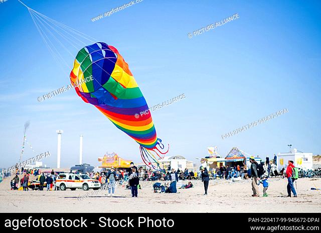 17 April 2022, Lower Saxony, Norderney: A colorful kite flies in the wind against a blue sky on Weststrand, while in the background numerous tourists watch the...