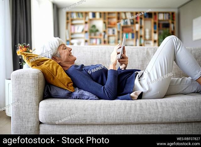Senior woman text messaging on digital tablet while lying down on sofa at home