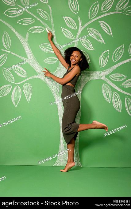 Smiling young woman standing on one leg drawing tree with chalk on green backdrop
