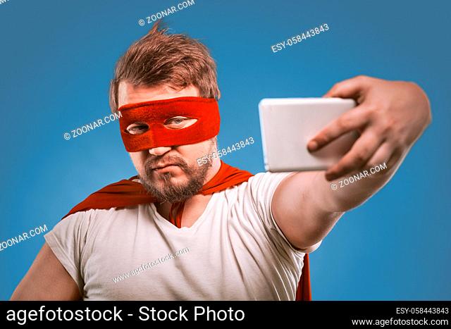 Super hero man takes selfie by mobile phones. Confident man in a super hero costume in a red mask and cloak poses on faded denim blue background making selfie...
