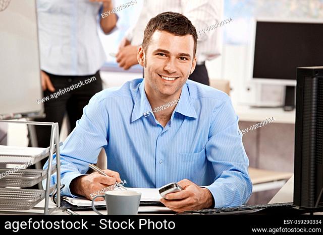 Casual young businessman working in office, sitting at desk, writing notes to personal organizer
