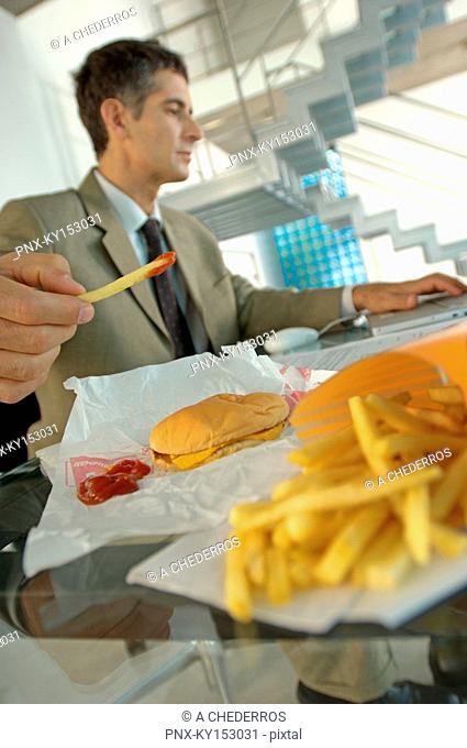 Mid adult businessman having burger and French fries, using laptop