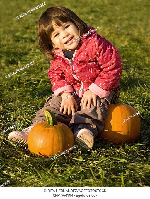 Smiley toddler sat on the green grass with 2 pumpkins