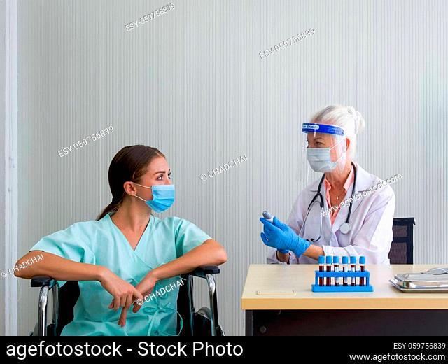An old woman doctor with a face shield and a protection mask inform the temperature measured result to the patient sitting in a wheelchair