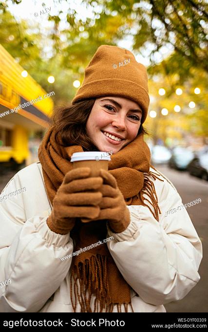 Smiling young woman wearing brown scarf with disposable coffee cup on street during winter