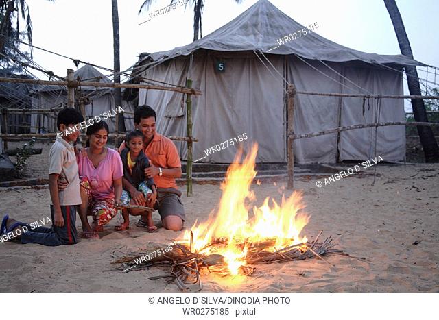 South Asian Indian parent and children sitting in front of campfire enjoying warmth in cold on seashore , Shiroda , Dist Sindhudurga , Maharashtra