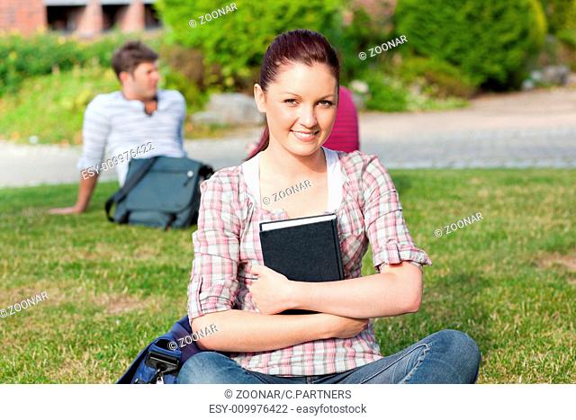 Smiling female student reading a book sitting on grass