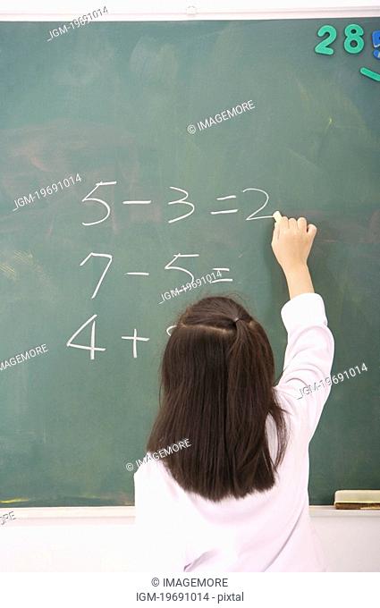 Girl solving mathematical equation on blackboard, rear view