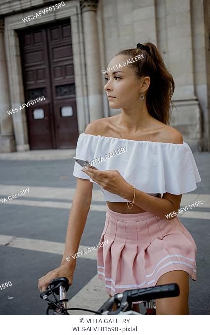 Fashionable young woman exploring Barcelona with a bicycle, using smartphone