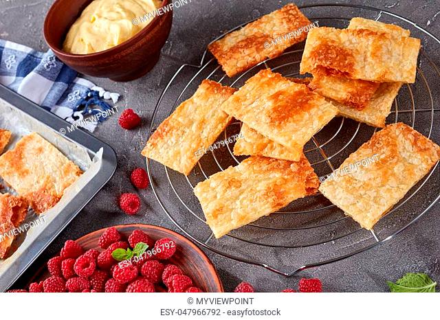 freshly baked portions of puff pastry on a baking sheet to cook french dessert millefeuille, fresh ripe raspberry on a plate and cream in a bowl on a concrete...