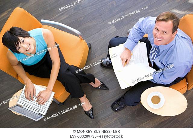 Two businesspeople sitting indoors with coffee laptop and folder smiling