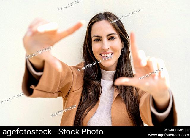 Smiling woman making picture frame with hands in front of white wall