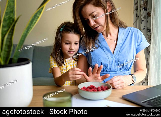 Smiling daughter putting raspberry on mothers finger at desk in home office
