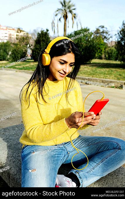 Smiling woman using mobile phone while listening music through headphones in public park
