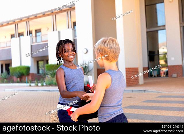 African american and caucasian elementary schoolboys with degree and mortatboard doing handshake