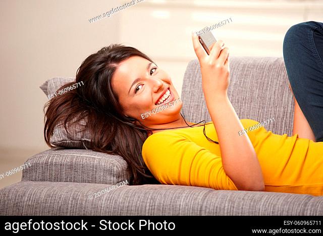 Happy teen girl holding phone with earphones lying on couch at home smiling looking at camera