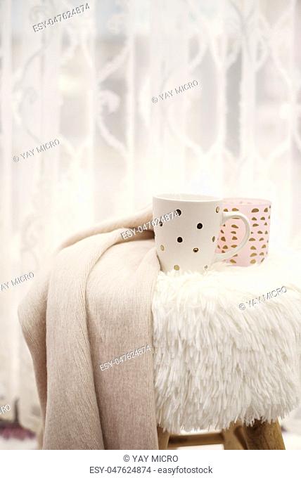 Hot chocolate, a cup of cappuccino on a fur chair in front of a large window with a white sheers curtain. Warm scarf and lights around