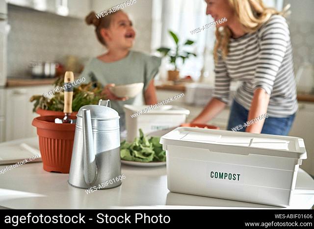 Compost container by watering can with woman and daughter in kitchen