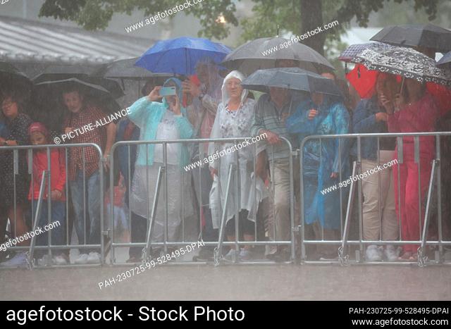 25 July 2023, Bavaria, Bayreuth: Onlookers stand outside the Festspielhaus on the Grüner Hügel in heavy rain before the opening of the Richard Wagner Festival...