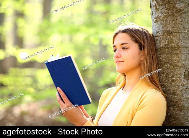Relaxed woman reading a paper book leaning on a tree in a beautiful forest