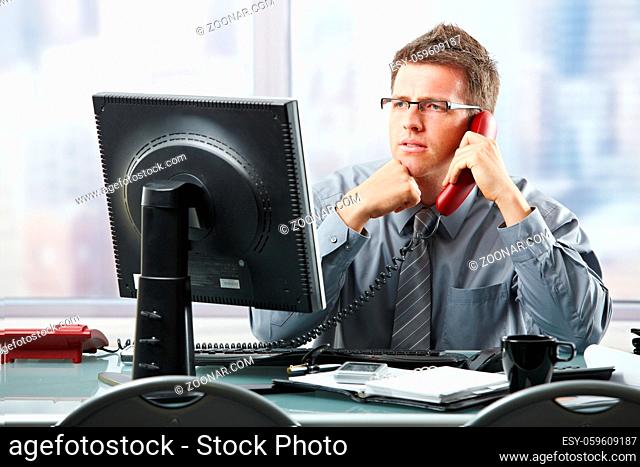 Focused businessman listening to explanation of computer report on landline phone looking at screen sitting in office