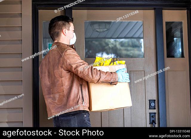A young man standing with a box of groceries at the door of a house during the Covid-19 World Pandemic; Edmonton, Alberta, Canada