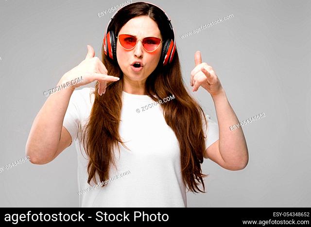 Brunette cheerful woman DJ in white T-shirt on and eyeglasses isolated on grey background with copyspace listen to music in red headphones, dancing and have fun