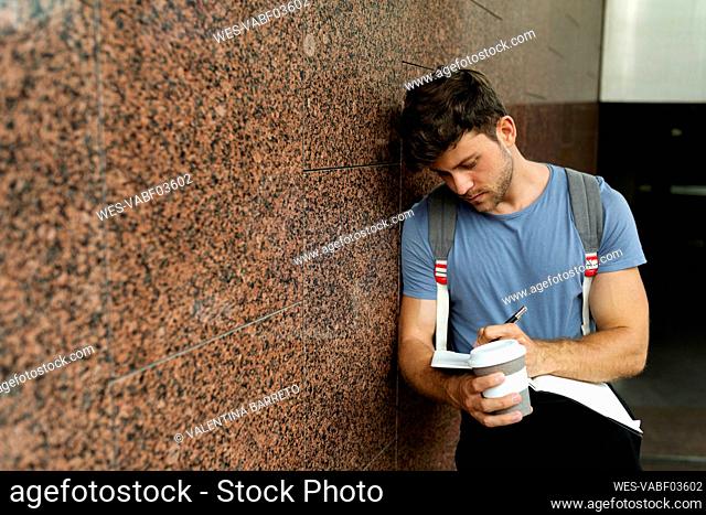 Handsome young man writing in book while holding coffee cup and leaning on brown tile wall at subway station