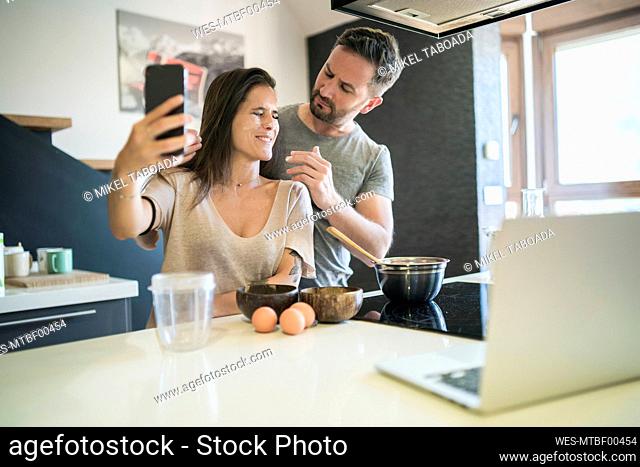 Playful man applying flour on woman's face while she is taking selfie with smart phone at home