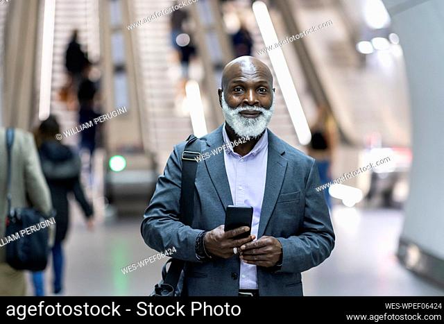 Smiling senior businessman with laptop bag and mobile phone at railroad station