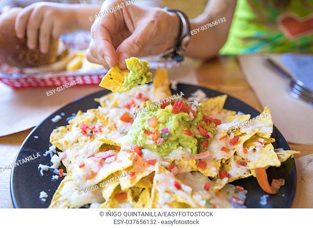 woman hand dipping nacho chips in avocado guacamole, cheese and chopped tomato on black plate, on wooden table with paper placemats at restaurant
