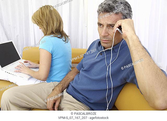Close-up of a mid adult man listening to an MP3 player with a mid adult woman using a laptop beside him