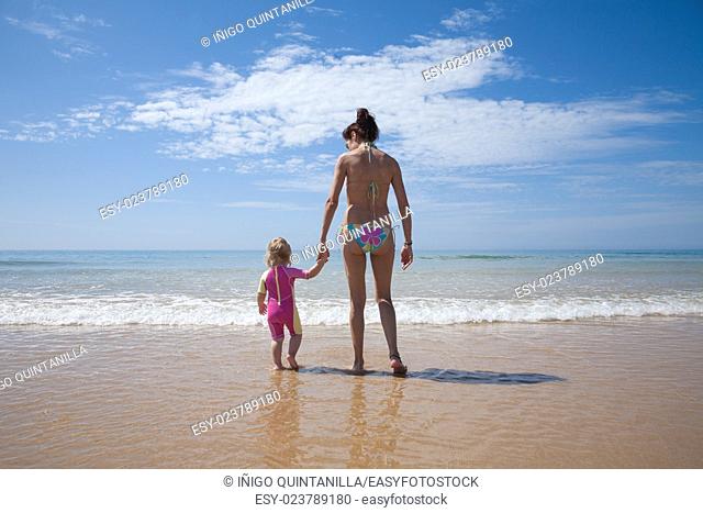 summer back family of two years old blonde baby with pink and yellow swimsuit holding hand with brunette woman mother in bikini standing at sea shore beach sand...